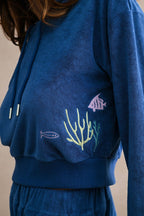 Close-up of blue terry cloth hoodie with embroidered fish and coral motifs on women's clothing.
