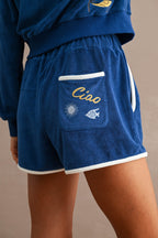 Close-up of blue terry cloth shorts with embroidered sun, fish, and “Ciao” text, featuring contrast white trim on a woman.