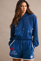 Woman wearing a cozy blue Ciao Terry Hoodie with matching shorts featuring colorful embroidered beach-themed designs, posing against a neutral background.