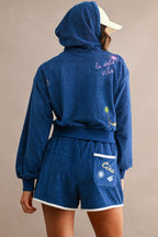 Woman wearing a navy blue embroidered terry cloth hoodie and shorts lounge set with beach-themed designs and contrast trim.