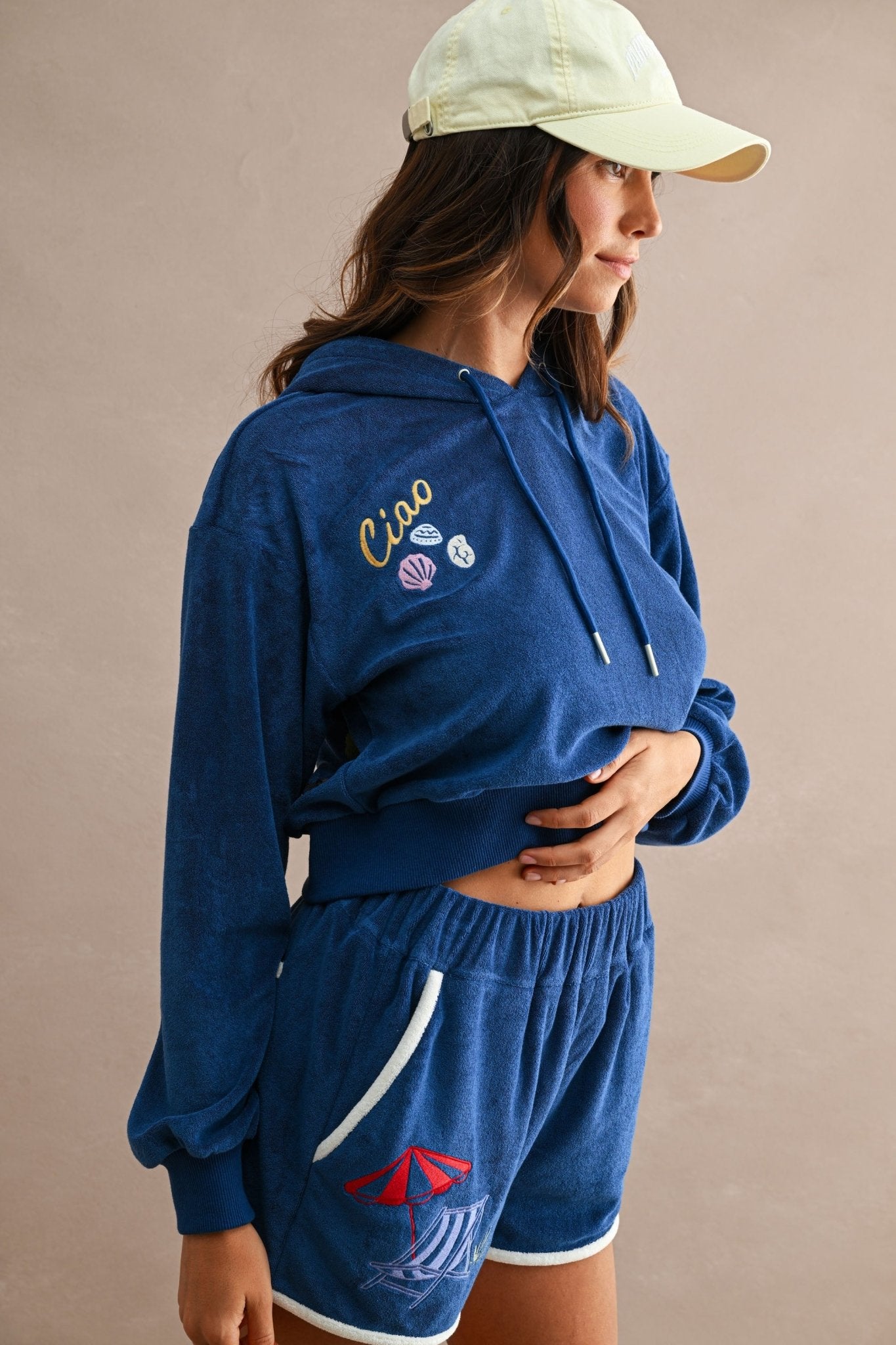 Woman wearing a navy blue Ciao Terry Hoodie with embroidered seashell and "Ciao" text, paired with matching shorts featuring a red umbrella and beach chair design, and a light-colored cap.