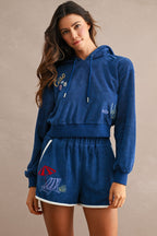 Woman wearing a blue terry cloth hoodie and matching Ciao Terry Shorts featuring embroidered beach-themed designs, including a red umbrella and lounge chair.