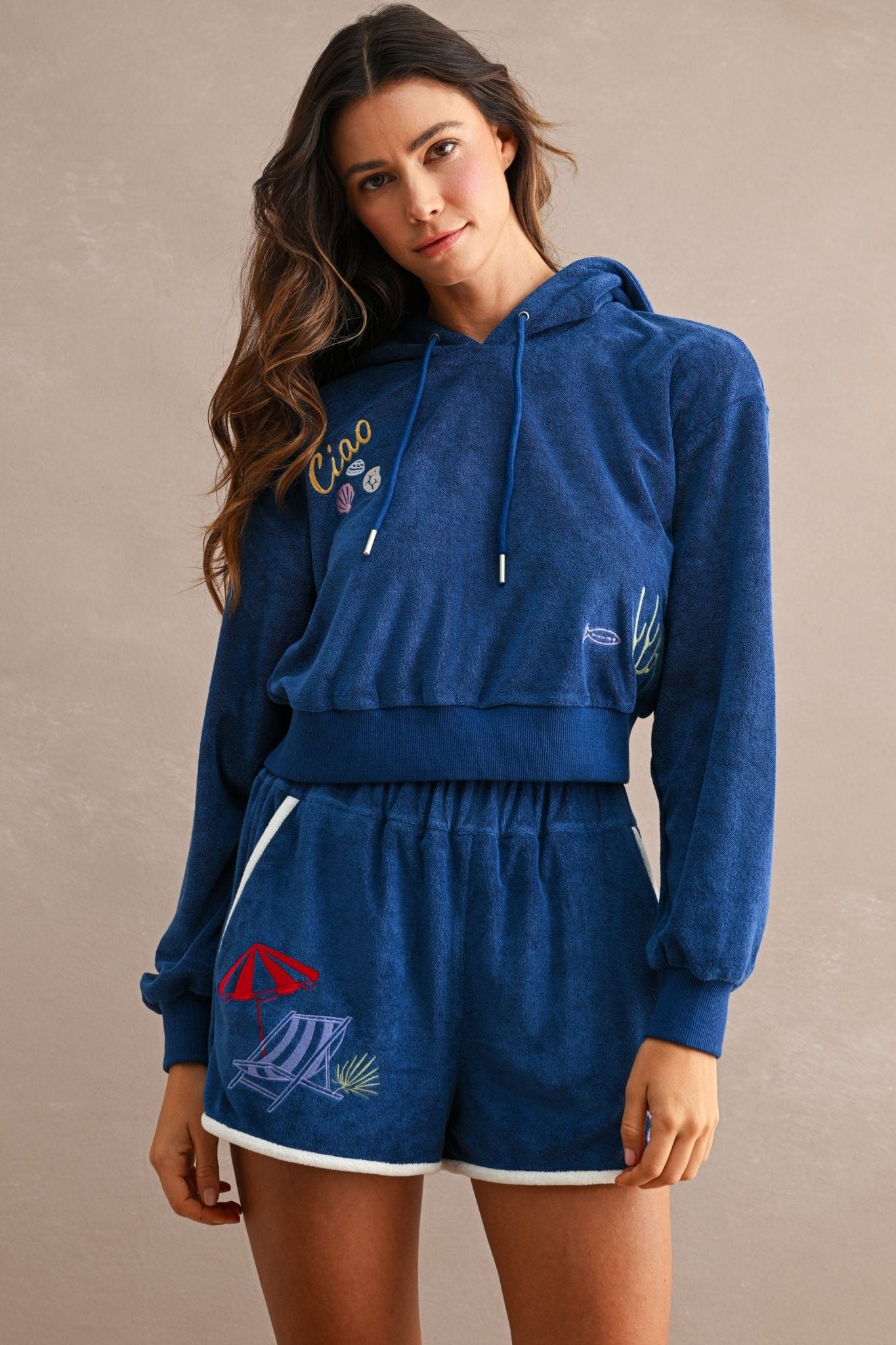 Woman wearing a blue terry cloth hoodie and matching Ciao Terry Shorts featuring embroidered beach-themed designs, including a red umbrella and lounge chair.