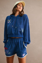 Woman wearing a blue terry cloth lounge set with embroidered details and contrast trim, styled with a beige cap.