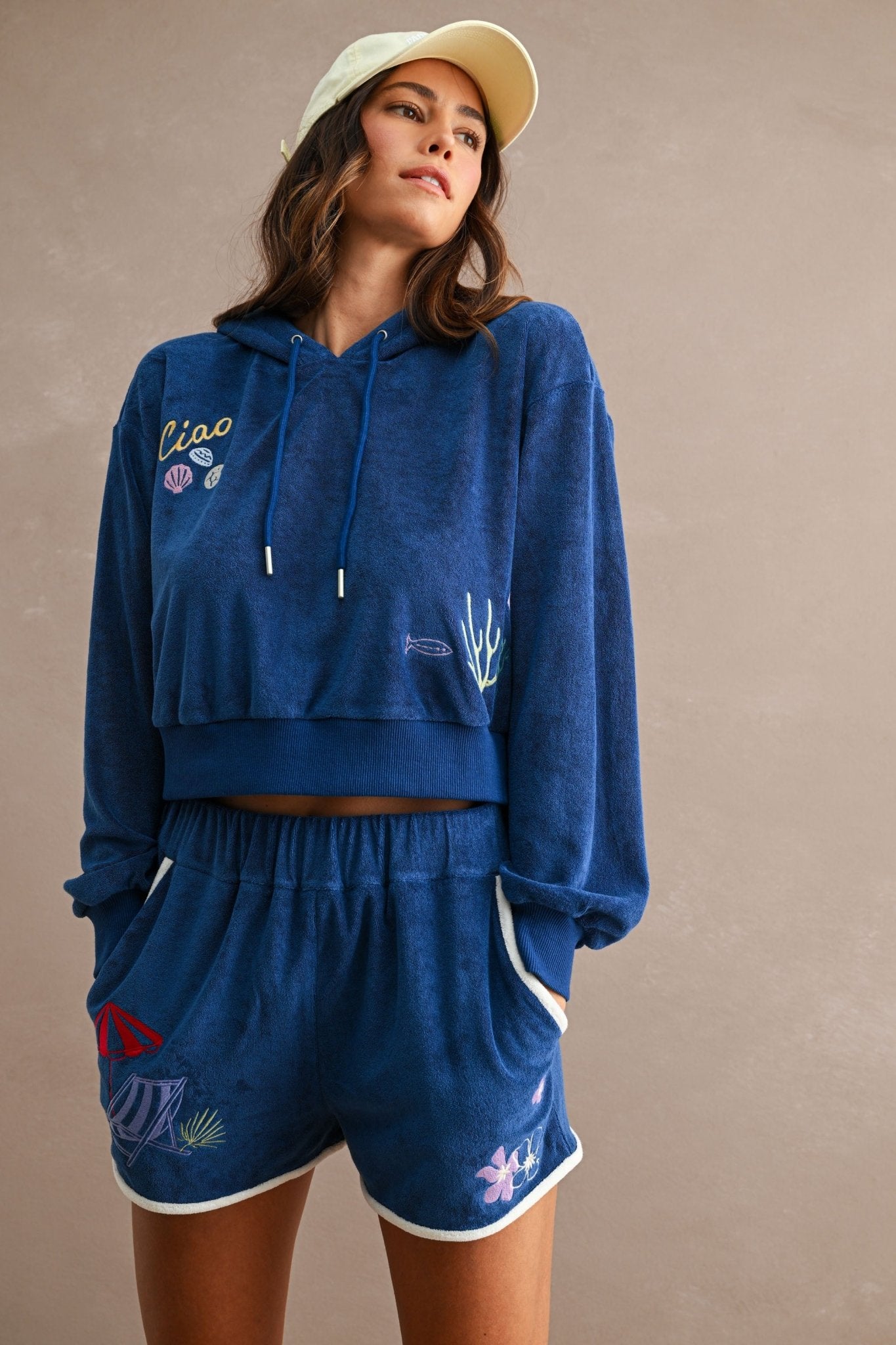 Woman wearing a blue hooded cropped sweatshirt with embroidered seashells and coral paired with matching blue Ciao Terry Shorts featuring white trim and embroidered beach motifs.