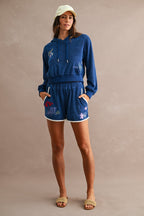 Woman wearing blue terry cloth Ciao Terry Shorts with embroidered beach motifs, paired with a matching hoodie, beige cap, and woven slide sandals.