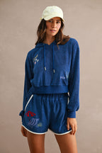 Model wearing navy blue terry cloth hoodie and Ciao Terry Shorts featuring a beach chair and umbrella embroidery, styled with a light yellow baseball cap.