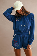 Woman wearing navy blue Ciao Terry Shorts with white trim and embroidered beach-themed designs, paired with matching long-sleeve top and light yellow cap.