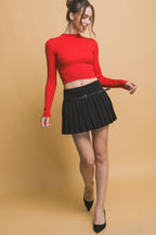 Woman wearing a red long-sleeve crop top and black pleated mini skirt with a slim belt in a studio setting