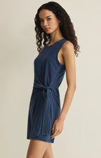 Woman wearing a sleeveless blue jersey denim mini dress with a side tie detail in a neutral setting
