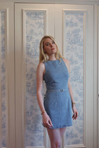 Woman wearing a fitted sleeveless denim mini dress with a belt, posing indoors against patterned wallpaper.