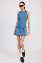 Woman wearing sleeveless denim mini dress with belt detail, paired with black shoes and white socks, posing on white background