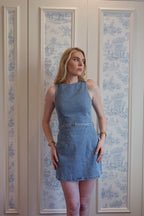 Woman wearing a sleeveless fitted blue denim mini dress standing against a patterned wall background