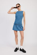 Woman wearing a sleeveless denim mini dress with belt, white socks, black loafers, and sunglasses posing against white background