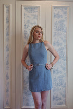 Woman wearing sleeveless denim mini dress with belt standing against white and blue patterned wall
