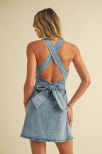 Back view of a woman wearing light blue denim mini dress with crisscross straps and tied bow at the waist