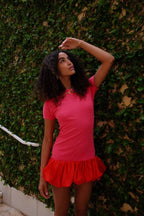 Woman wearing a fitted pink mini dress with a ruffled red hem standing against a green ivy wall outdoors