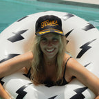 Woman wearing black embroidered trucker hat with "Tequila" text, smiling in a pool on a white lightning bolt float.