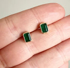 Small rectangular green gemstone stud earrings with gold setting held on fingers against neutral background