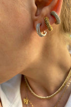 Close-up of a woman's ear wearing three textured silver and gold hoop earrings, paired with layered gold necklaces.