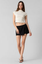 Woman wearing a cream crop top and black tailored skort standing against a plain gray background.