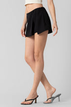 Woman wearing black tailored skort paired with beige crop top and black heeled sandals on gray background