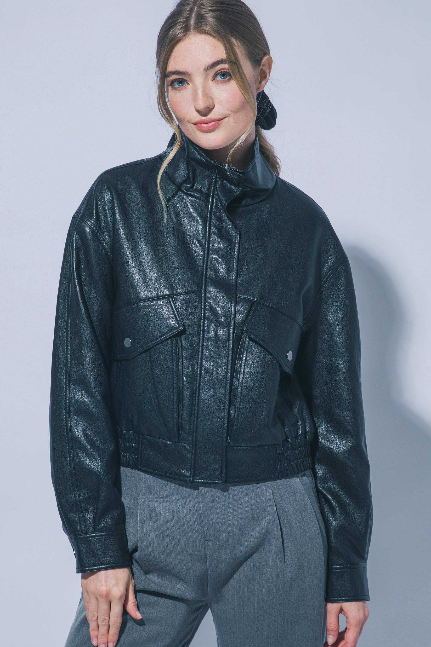 Woman wearing a black faux leather jacket with large front pockets and a high funnel collar in a studio setting