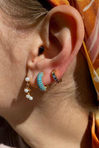 Close-up of an ear with gold hoop earrings featuring tiny dangling pearls and turquoise and rainbow accents.