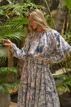 Woman wearing a flowing bohemian maxi dress with blue nature-inspired prints and a decorative belt in a tropical garden setting