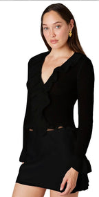Woman wearing a black knit cardigan with ruffled neckline and long sleeves paired with a black skirt.