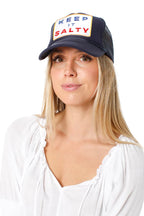 Woman wearing navy foam front trucker hat with "KEEP IT SALTY" patch and white blouse in women's clothing style.