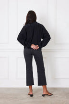 Woman wearing a black shirt with billowy sleeves and dark cropped jeans standing against white wall