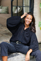 Woman wearing a black blouse with billowy sleeves and embroidery, smiling and sitting outdoors in casual women's clothing.