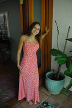 Woman wearing a pink floral maxi dress with adjustable straps, standing indoors near a wooden door and plant pot.