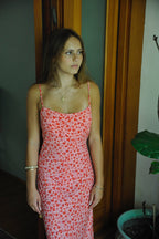 Young woman in a red floral maxi dress with thin straps standing indoors near wooden doors and a plant.
