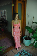 Woman wearing pink floral maxi dress with thin straps standing barefoot indoors near potted plant and wooden door