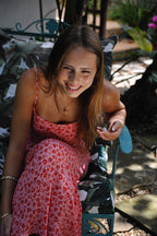 Smiling woman in a pink floral maxi dress sitting outdoors on a patterned cushion chair in natural light