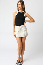 Woman wearing a black sleeveless ringer tank top with white trim paired with a beige mini skirt with pockets.