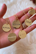 Hand-stamped gold-tone pendant necklaces with casual phrases on delicate chains, displayed on a hand with rings.