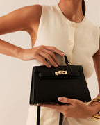 Woman holding black leather handbag with gold clasp, wearing sleeveless cream buttoned top and gold bracelets