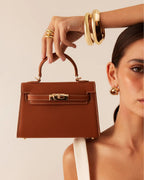 Woman holding a brown leather handbag with gold hardware, wearing gold bracelets and earrings, fashion accessory detail