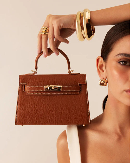 Woman holding a brown leather handbag with gold hardware, wearing gold bracelets and earrings, fashion accessory detail