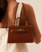 Brown leather handbag with gold hardware and top handle worn over shoulder by woman in white dress
