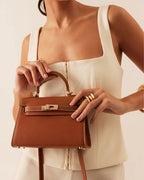 Woman wearing cream top holding brown leather handle bag with gold hardware and stitching details