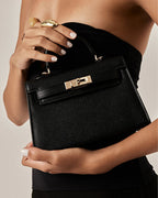 Woman holding a black textured leather handle bag with gold hardware and a clasp closure, wearing a strapless black top