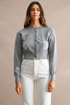 Woman wearing a grey embroidered cardigan with button front and white high-waisted pants against neutral background.