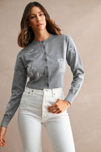 Woman wearing a grey embroidered cardigan with white floral and animal designs paired with white pants.
