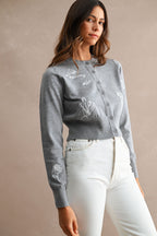 Woman wearing a gray embroidered button-up cardigan paired with high-waisted white jeans in a neutral setting.