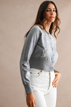 Woman wearing a gray embroidered cardigan with floral designs paired with white pants, modeling women's clothing.