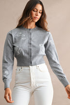 Woman wearing a gray embroidered cardigan with floral and animal designs paired with high-waisted white pants.